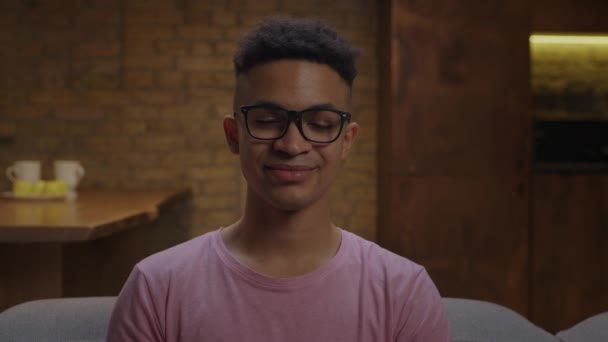 Pretty young African American man wearing eyeglasses biting off apple looking at camera sitting on couch at home. Handsome man chewing apple fruit and smiling. — Stock Video