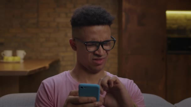 Verrast jonge Afro-Amerikaanse man scrollen mobiele telefoon scherm met vingers en denken zitten op de bank. close-up van de mens het maken van ontevreden gezicht. — Stockvideo