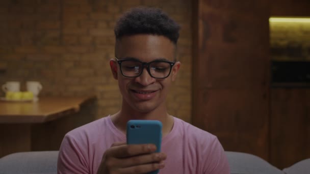Joven hombre afroamericano con anteojos usando el teléfono móvil y navegar en línea sentado en el sofá en casa. Primer plano de gadget adicto millennial masculino de navegación en la red social. — Vídeos de Stock