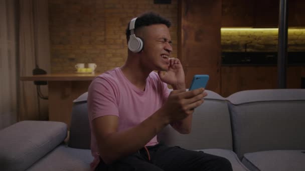 Millennial afroamerikansk man i hörlurar sjunger karaokesång tittar på smartphone sitter på soffan hemma. Positiv ung vuxen man tycker om att sjunga ensam. — Stockvideo