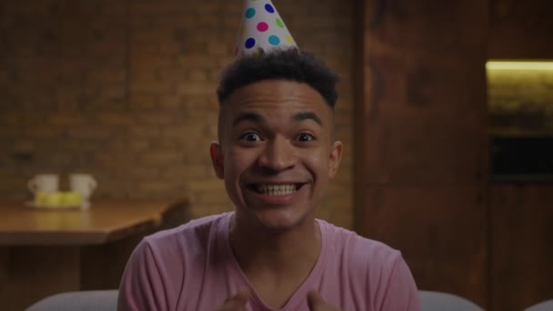 Jeune homme afro-américain adulte portant un chapeau d'anniversaire chantant une chanson d'anniversaire à la caméra. Webcam vue de l'homme félicitant quelqu'un en ligne. — Video