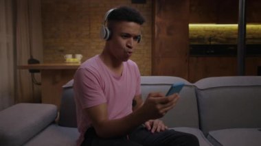 20 'li yaşlarda, kablosuz kulaklık takan, elinde cep telefonuyla kanepede oturan Afro-Amerikan bir adam şarkı söylüyor. Neşeli genç yetişkin adam evde şarkı söyleyip dans etmekten zevk alıyor..