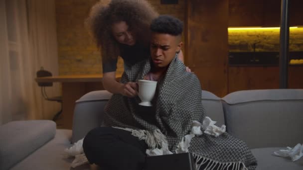 Caring giovane moglie adulta dando bevanda tè caldo al marito afroamericano malato. Razza mista coppia familiare sensazione di malessere malattia seduta sul divano a casa. — Video Stock