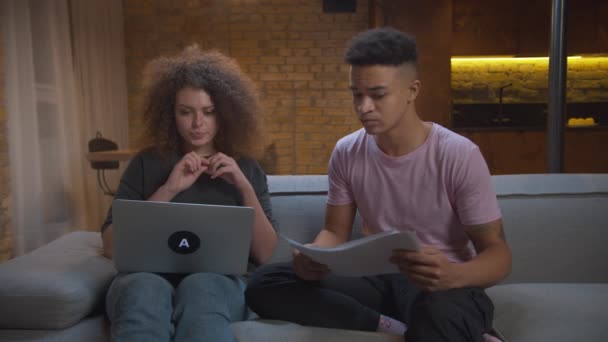 Millennial multiethnischen Paar betrügen, während Online-Interviews für einen neuen Job. Afroamerikanisch Freund hilft Freundin, Online-Interview Fragen zu beantworten sitzt auf der Couch zu Hause. — Stockvideo