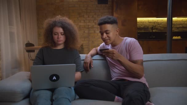 20 대 다양 한 민족들이 집에서 온라인으로 물건을 사는 것을 행복하게 생각 합니다. 온라인 쇼핑을 위해 컴퓨터를 이용하는 아프리카계 미국인 남자와 여자가 소파에 앉아 있습니다. 가족의 하이파이브 !. — 비디오