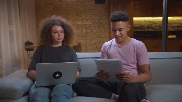 Afro-Amerikaanse student uit de 20 helpt zijn vriendin met huiswerk op laptop. Jonge volwassen man en vrouw samen online studeren thuis zitten op de bank. — Stockvideo