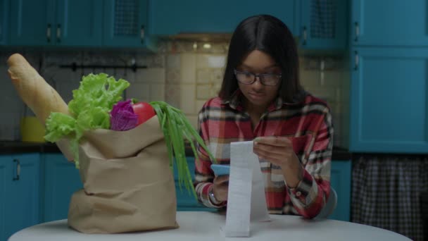 20-talets afroamerikanska kvinna kollar maten i kassen med sitt matkvitto, kostnadsspårning. Kvinnliga inspektera stormarknad faktura med mobiltelefon. — Stockvideo