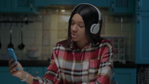 Young African American woman singing songs holding mobile phone in hand wearing white headphones. Millennial woman enjoys singing with cell phone and dancing at blue kitchen at home. — Stock Video