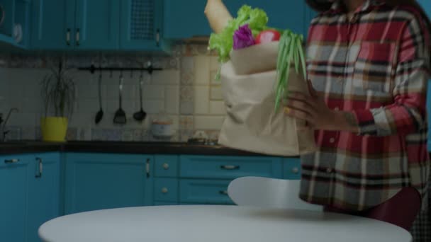 Jovem mulher afro-americana adulta em óculos cansados de segurar saco de papel com comida fresca sentada na mesa de jantar na cozinha azul em casa. — Vídeo de Stock