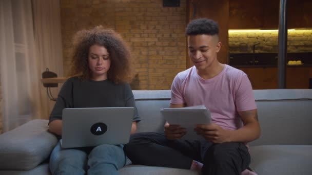 Pareja joven de raza mixta estudiando juntos en línea sentados en el sofá en casa. Afro-americano estudiante masculino leyendo documentos en papel y dictando a la mujer escribiendo en el portátil. — Vídeos de Stock