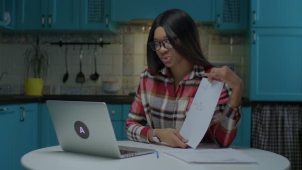 20s profesor afroamericano en gafas hablando y mostrando hoja de papel con ecuaciones físicas mirando portátil. Trabajo desde casa profesor de escuela en línea. — Vídeo de stock