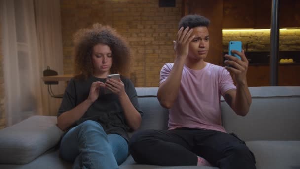 Pareja joven de raza mixta usando gadgets sentados en el sofá en casa. Afro-americano hombre hablando con el teléfono móvil de la cámara y la mujer navegar por Internet. — Vídeos de Stock