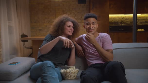 20s woman checking mobile phone notifications while watching TV with African American boyfriend sitting on couch at home. Millennial mixed race couple eating pop corn in front of tv. — Stock Video