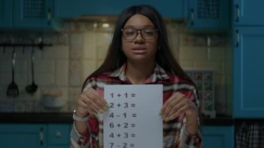 Gözlüklü, genç, Afro-Amerikan bir bayan öğretmen kamerayla konuşuyor ve basit matematiksel denklemlerle kağıdını gösteriyor. Web kamerası görüntüsü evde online öğretmenlik yapıyor..