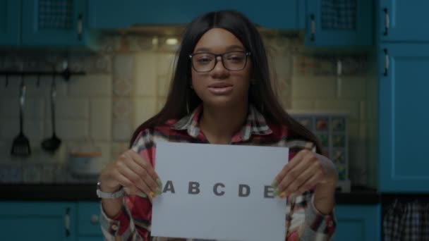 Vrouwelijke Afro-Amerikaanse leraar in bril in gesprek met camera en het tonen van vel papier met ABCDE letters. Webcam weergave van werken vanuit huis online school leraar. — Stockvideo