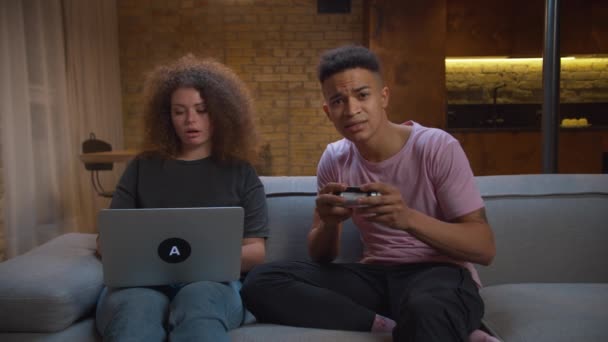 20-as évekbeli afro-amerikai férfi videojátékozik dühös feleséggel, aki otthon laptopozik. Férj zavaró feleség dolgozik online ül a kanapén. — Stock videók