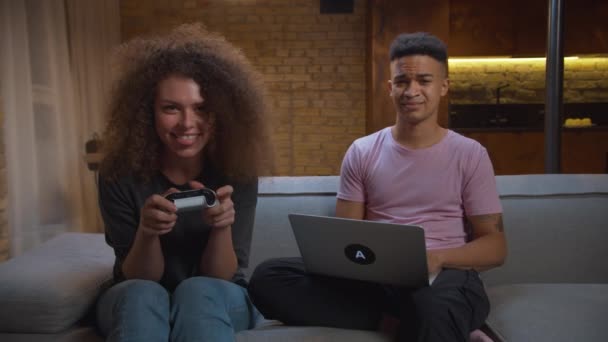 Mujer milenaria jugando videojuegos con su novio afroamericano trabajando en la computadora portátil en casa. Jóvenes de raza mixta de la familia pasar la noche juntos en el sofá. Esposa perturba a marido trabajador. — Vídeo de stock