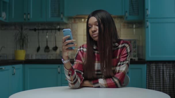年轻的非洲裔美国女人坐在厨房桌子旁，一边用手机一边上网。Gadget上瘾的千年女性浏览社交网络. — 图库视频影像