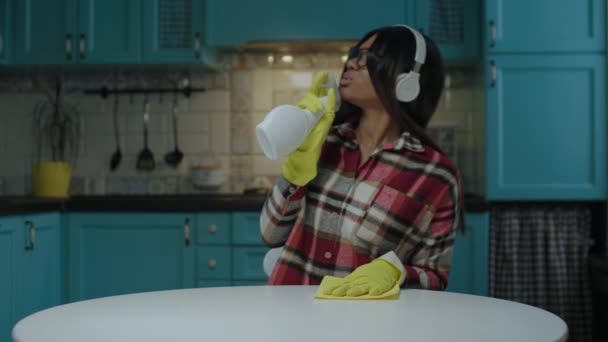 Jonge volwassen Afro-Amerikaanse vrouw luisteren muziek in hoofdtelefoon tijdens het schoonmaken tafel met schoonmaakmiddel en doek dragen van gele rubberen handschoenen in de blauwe keuken. — Stockvideo