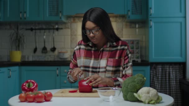 Joven mujer afroamericana adulta cocinando ensalada de verduras frescas sentada en la mesa de comedor en la cocina. — Vídeo de stock