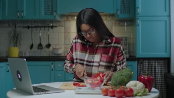Молодая афроамериканка учится готовить, используя кухонные весы и глядя на онлайн рецепт на ноутбуке, сидя за кухонным столом. 20-летняя женщина готовит свежий овощной салат глядя на ноутбук. — стоковое видео