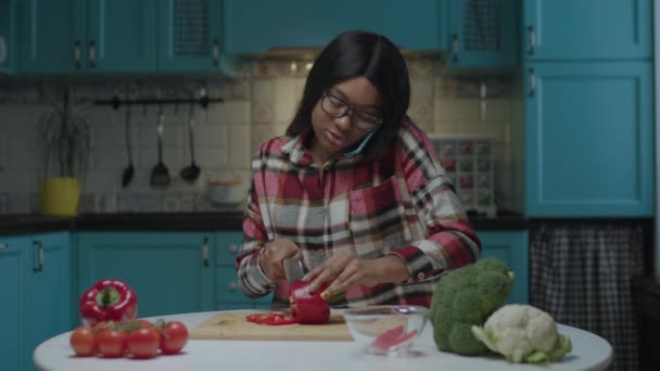 20s Afro-Amerikaanse vrouw praten door mobiele telefoon en het koken van verse groente salade zitten aan de eettafel op de keuken. — Stockvideo