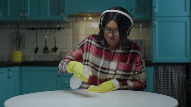 Millennial mulher afro-americana cantando enquanto limpava a mesa com agente de limpeza e pano usando luvas de borracha amarela e fones de ouvido sem fio na cozinha. — Vídeo de Stock