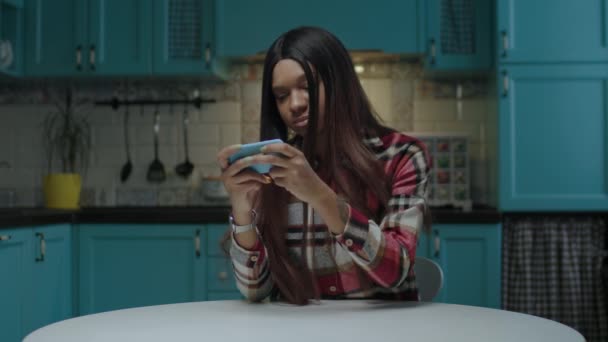 Fiatal afro-amerikai nő videojátékozik mobiltelefonnal a konyhában. 20-as évek nő mobiltelefon játék online játékok. — Stock videók