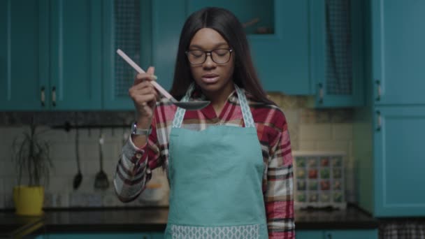 Mujer afroamericana bonita en delantal sosteniendo cucharada y degustación de comida mirando a la cámara. Cocinera de pie en cocina azul. — Vídeos de Stock