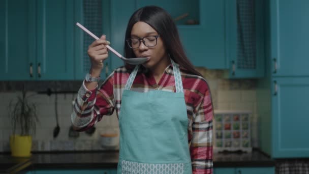 年轻的非洲裔美国女厨师穿着围裙，用勺子在镜头前品尝食物。20多岁的女人喜欢在家里做饭. — 图库视频影像