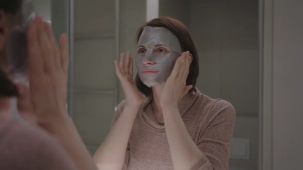 Młoda, dorosła kobieta w masce kosmetycznej i podziwiająca z lustrem w łazience. Happy kobieta podejmowania domu zabiegi kosmetyczne spa. — Wideo stockowe