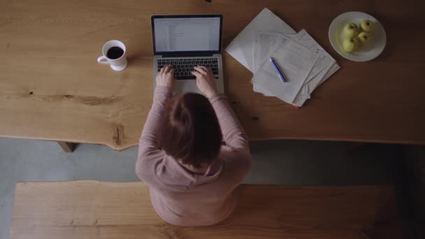 Draufsicht einer Frau in den 30er Jahren, die auf einem Laptop mit Papierdokumenten am Schreibtisch sitzt. Junge erwachsene weibliche Fernarbeit oder Fernlernen von zu Hause aus. — Stockvideo