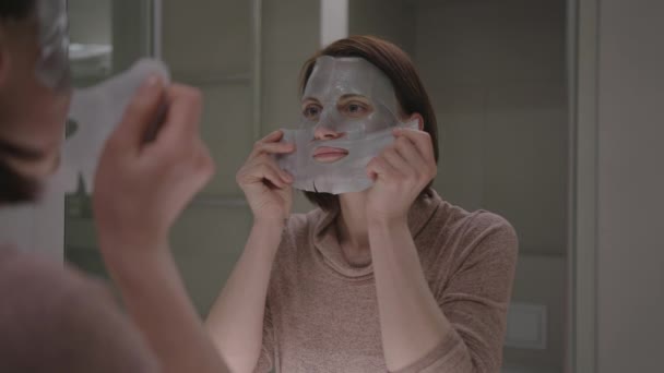 Молодая взрослая женщина делает домашние процедуры красоты стоя в ванной комнате. Женщина 30-х годов надевает маску для лица дома. — стоковое видео
