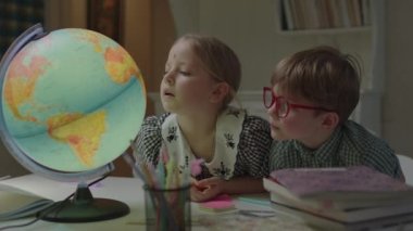 İki çocuk okul kitaplarıyla birlikte masada Dünya küresi üzerinde çalışıyor. Liseli kız ve anaokulu çocuğu evde birlikte öğreniyorlar..