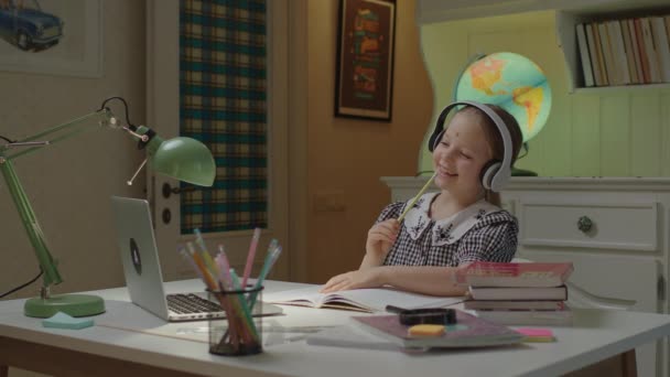Glad skolflicka sjunger med blyertspenna som mikrofon och tittar på laptop. Ungen bär trådlösa hörlurar lyssnar på musik och sång på nätet. — Stockvideo