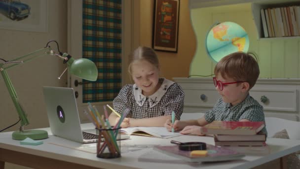 Zwei Kinder schreiben in Notizbüchern während Fernunterricht und Online-Unterricht am Laptop. Schulmädchen und Vorschulkind lernen zu Hause gemeinsam per Internet. — Stockvideo