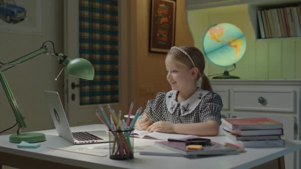 Kindergroet online leraar en opschrijven in notebook met pen op zoek naar laptop. Schoolmeisje leren online thuis. Leren op afstand voor kinderen. — Stockvideo