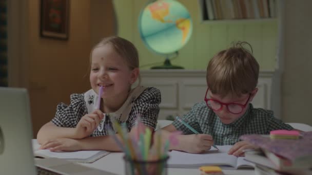 Dos niños que aprenden a distancia en casa escuchando al profesor en línea en el portátil sentado en el escritorio con cuadernos. Hermanos escribiendo tareas para la escuela de internet. — Vídeos de Stock