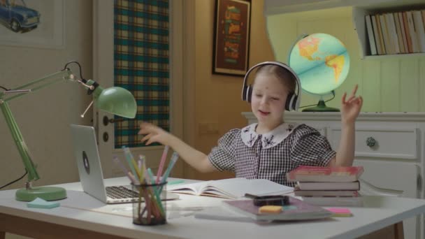 Mooi schoolmeisje dat naar moderne muziek luistert en thuis aan het bureau zit te dansen. Afstandsonderwijs en thuisonderwijs voor kinderen. — Stockvideo