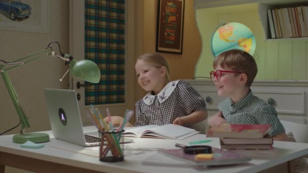 Two siblings have online lesson sitting at desk at home. Schoolchildren making fun during home schooling. Brother and sister laughing looking at laptop. — Stock Video