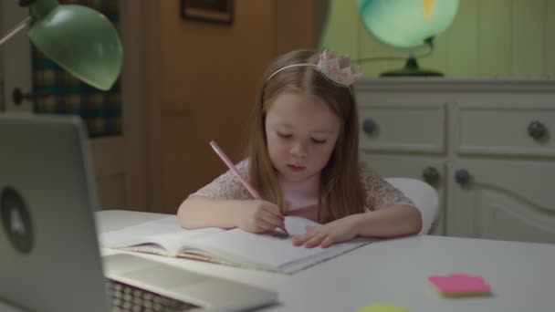 Nahaufnahme eines niedlichen Vorschulmädchens, das mit Bleistift in ein Notizbuch schreibt und auf den Laptop schaut. Das weibliche Kind lernt zu Hause online. Fernlernen für Kinder. — Stockvideo