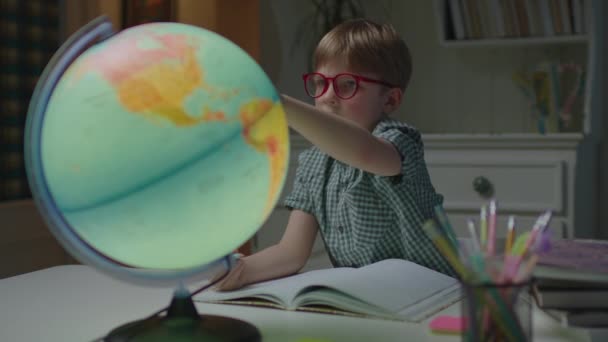 Vorschulkind studiert Erdkugel auf dem Tisch mit Schulbüchern. Schuljunge mit Brille lernt zu Hause. — Stockvideo