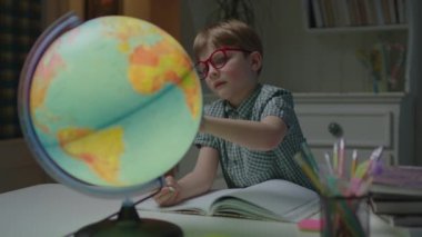 Anaokulu çocuğu evde coğrafya okuyor. Elinde defterle masada Dünya küresi kullanıyor. Gözlük takan okul çocuğu evde tek başına öğreniyor..