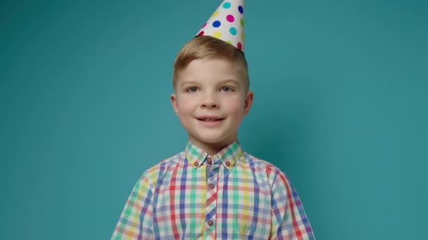 Niño joven con sombrero de cumpleaños felicitando a hablar con la cámara en el fondo azul. Alegre niño preescolar cantando canción de cumpleaños. — Vídeos de Stock