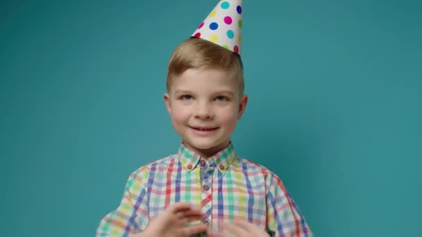 Doğum günü partisi şapkalı, gülümseyen anaokulu çocuğu el sallıyor ve mavi arka planda kamerayla konuşuyor. Mutlu çocuk doğum gününü kameraya bakarak kutluyor.. — Stok video