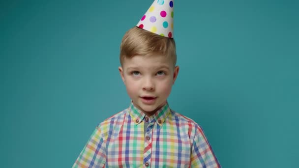 Aranyos fiú szülinapi sapkában, kék háttérrel beszél a kamerába. Webkamera nézet a gyerekek online születésnapi üdvözlet. — Stock videók