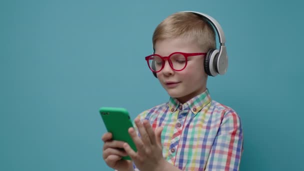 Miúdo a ver o telemóvel a segurar as mãos e a dar telemóvel a alguém. Menino em óculos e fones de ouvido usando o aplicativo de telefone inteligente no fundo azul. — Vídeo de Stock