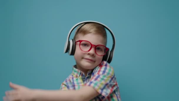 Preschool boy wearing eye glasses and wireless headphones enjoying music on blue background smiling and looking at camera in slow motion. — Stock Video