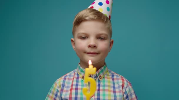 Niño en sombrero de cumpleaños soplando la vela número 5 en cámara lenta. Un niño de cinco años celebra su cumpleaños. Sonriente cumpleañero sobre fondo azul. — Vídeos de Stock