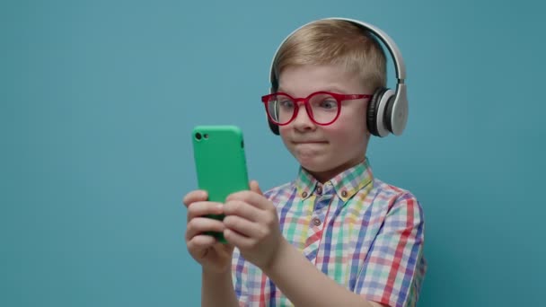 El niño preescolar salió usando un teléfono móvil con auriculares inalámbricos y anteojos. Niño sosteniendo el teléfono inteligente en la mano sobre fondo azul. — Vídeo de stock